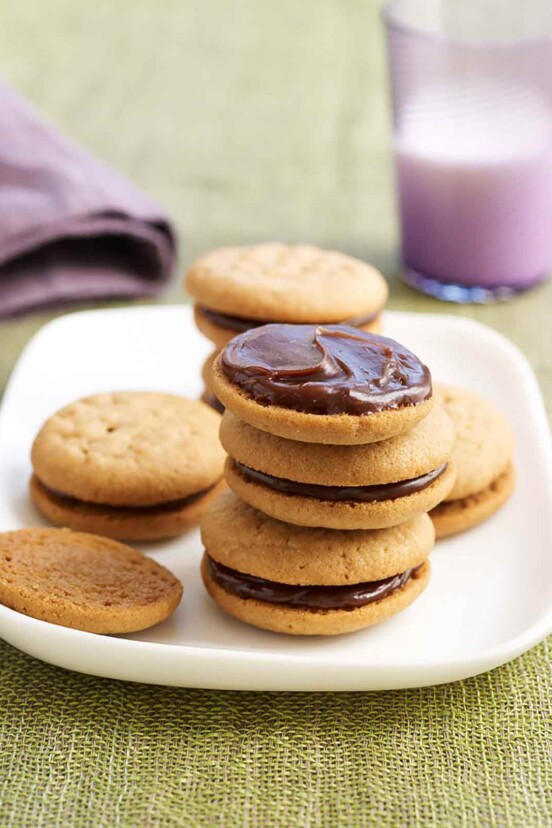 Peanut butter sandwich cookies stacked on a plate, the top one is without its top cookie. A glass of milk is in the background.