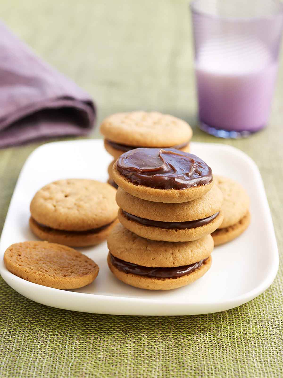 Peanut butter sandwich cookies stacked on a plate, the top one is without its top cookie. A glass of milk is in the background.