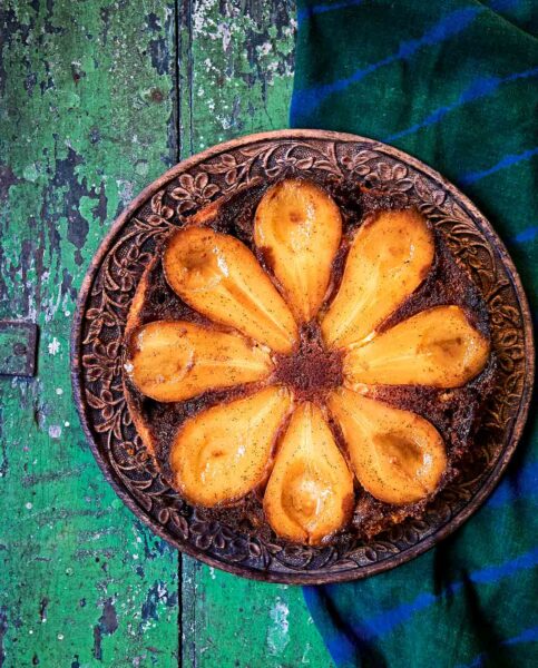 A pear and cardamom caramel upside-down cake on a carved wooden plate, on a green wooden table.