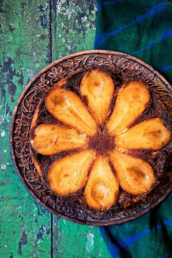 A pear and cardamom caramel upside-down cake on a carved wooden plate, on a green wooden table.