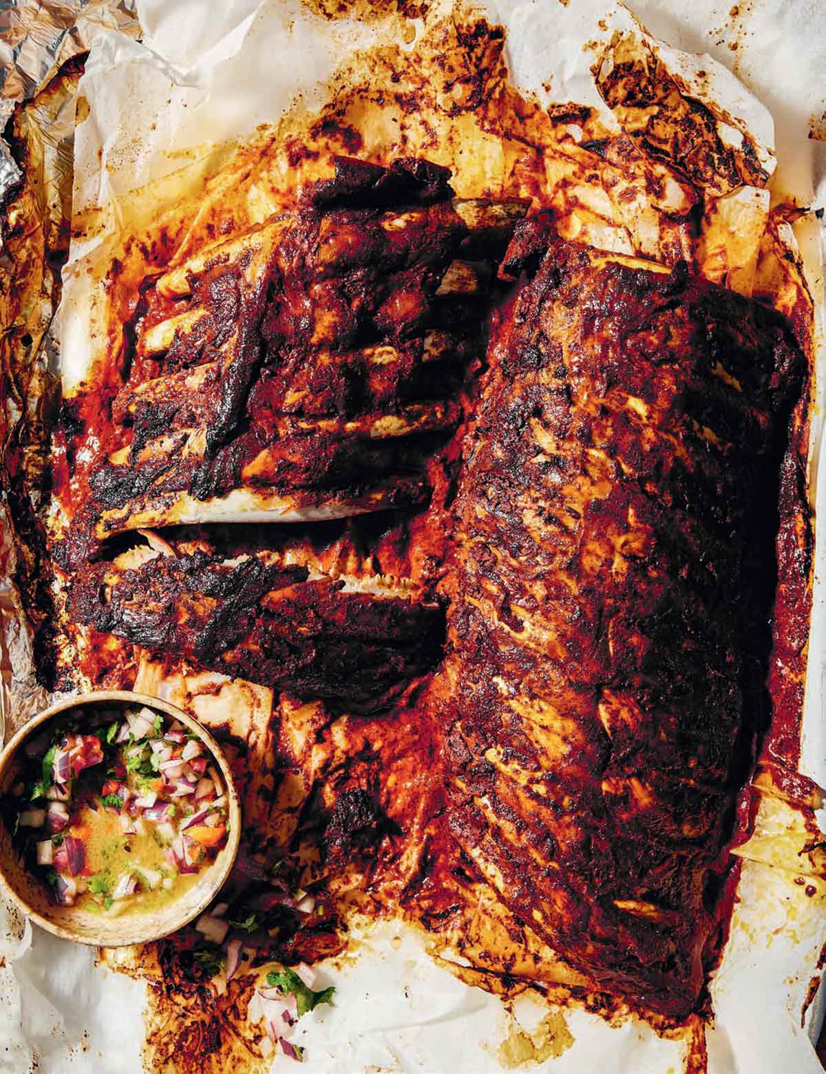 Pibil-style pork ribs on sheets of parchment paper with a bowl of habanero salsa next to it.