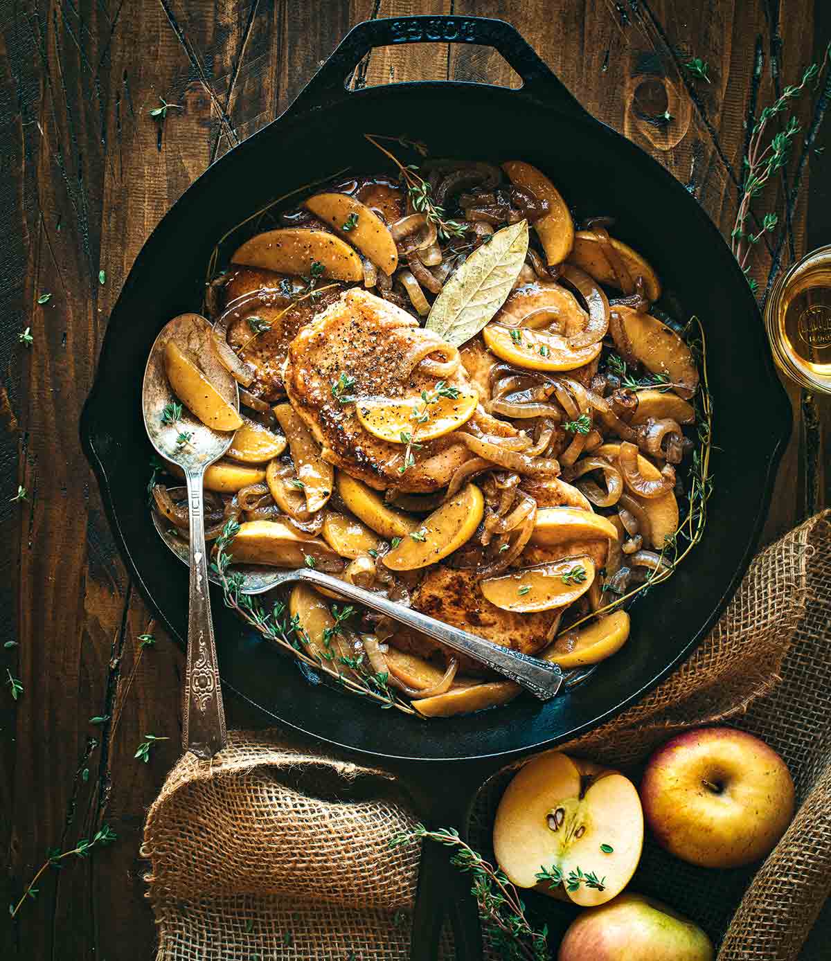 Paleo pork Normandy garnished with thyme, in a cast-iron pan with two serving forks, surrounded by burlap, apples, and thyme.