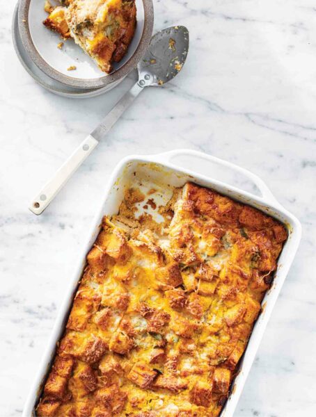 Pumpkin, roasted squash, and Gruyère strata in a white casserole dish with one piece in a small bowl, next to a metal serving spoon.