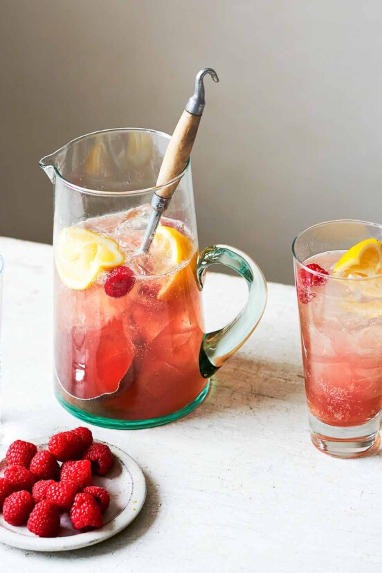 Raspberry vodka lime soda in a pitcher with a large spoon, beside two full glasses, one empty glass, and and a dish of raspberries.