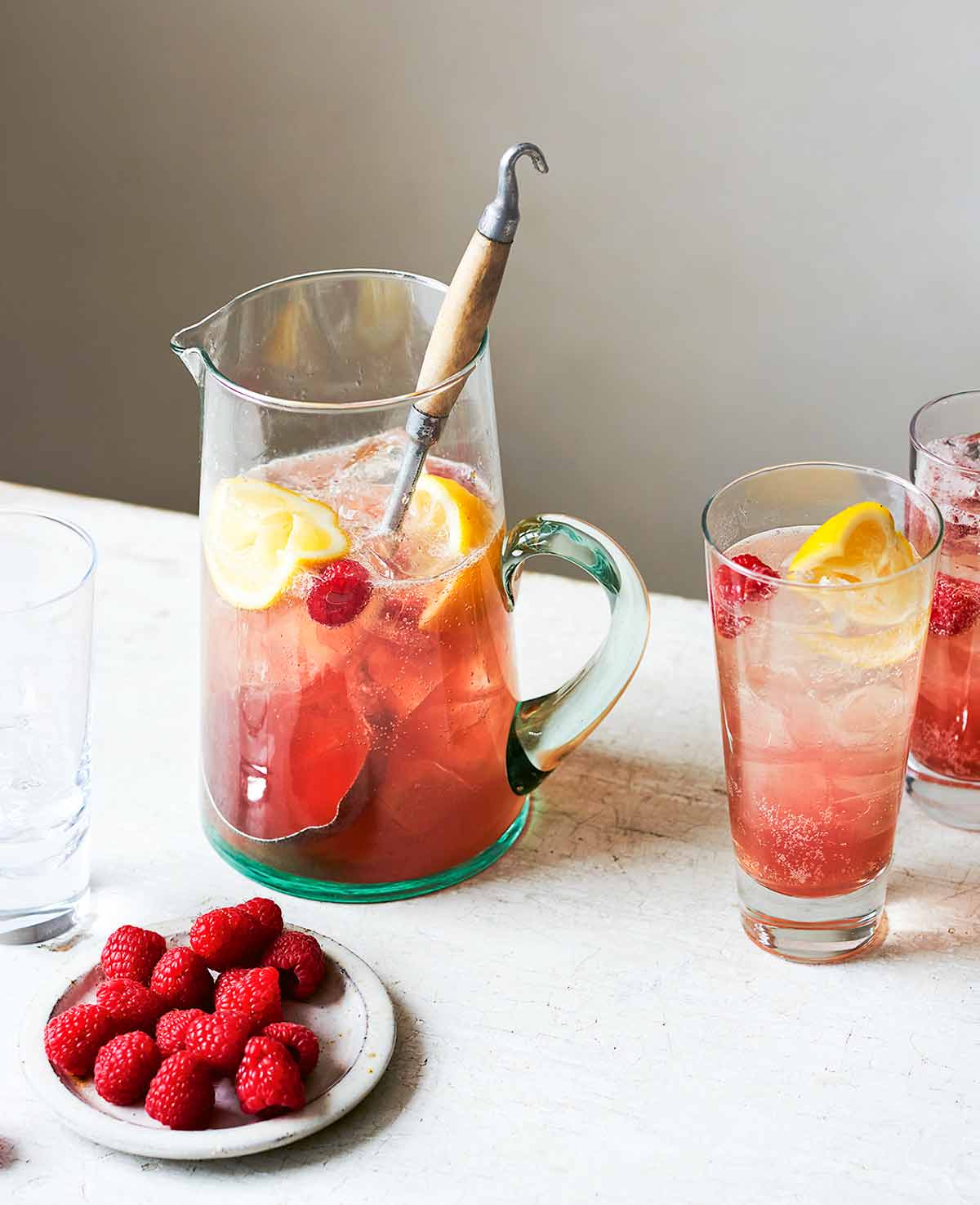 Raspberry vodka lime soda in a pitcher with a large spoon, beside two full glasses, one empty glass, and and a dish of raspberries.