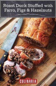 A roast duck stuffed with farro, figs, and hazelnuts, with three slices cut from it on a wooden cutting board with a knife resting beside it.