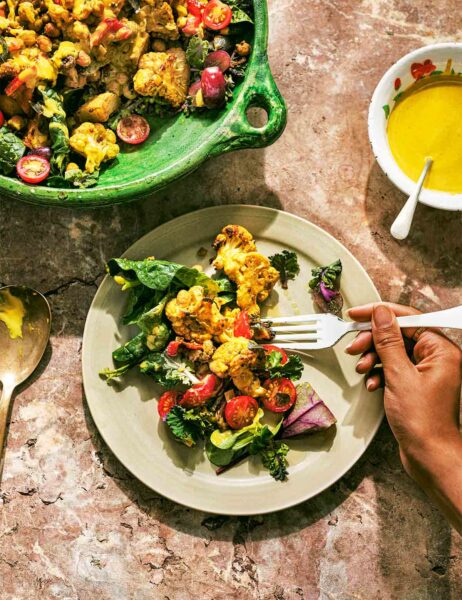 Roasted cauliflower curry on a plate with a hand holding a fork, beside a large pot filled with curry and a small bowl of curry sauce.