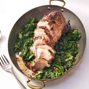 Roasted pork shoulder with mustard glaze on a bed of wilted mustard greens with sweet potatoes and a serving fork.