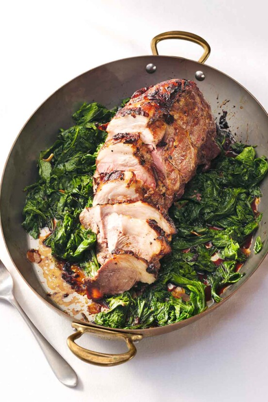 Roasted pork shoulder with mustard glaze on a bed of wilted mustard greens with sweet potatoes and a serving fork.