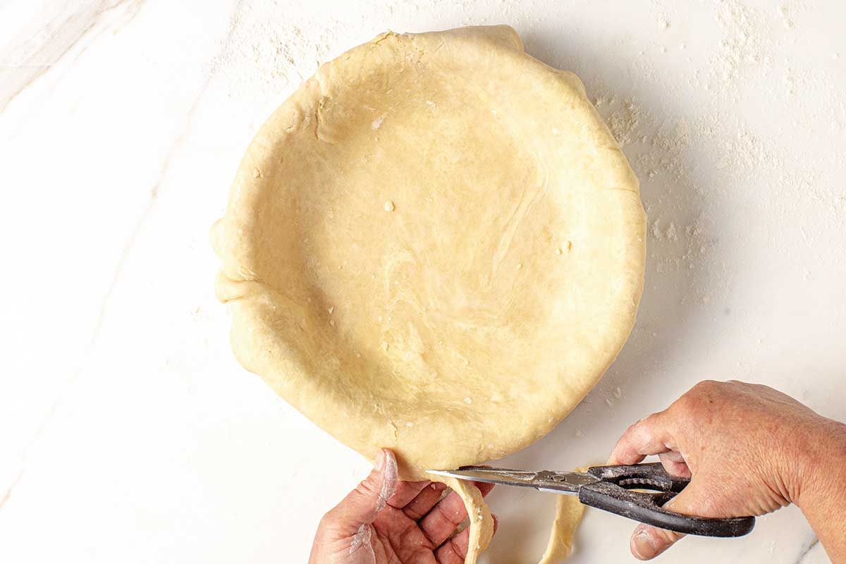 A person trimming a rolled pie crust with scissors.