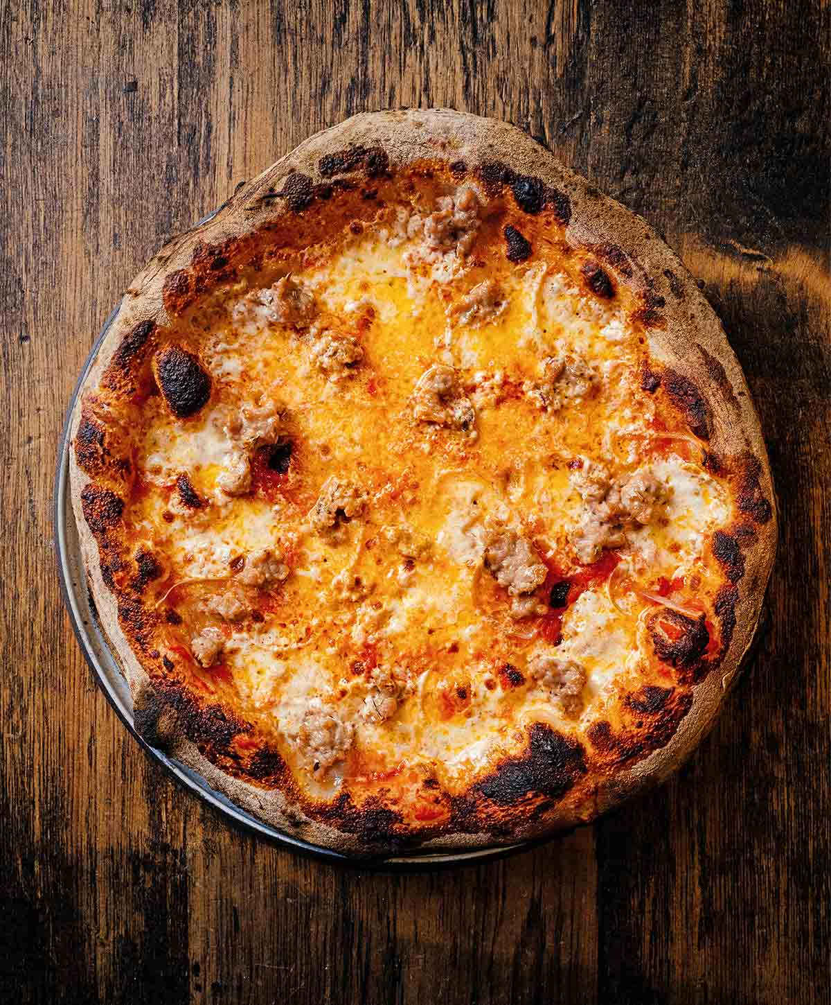 Sausage pizza with mozzarella and chili oil with a bubbled and blisters top, in a pizza pan on a wooden table.