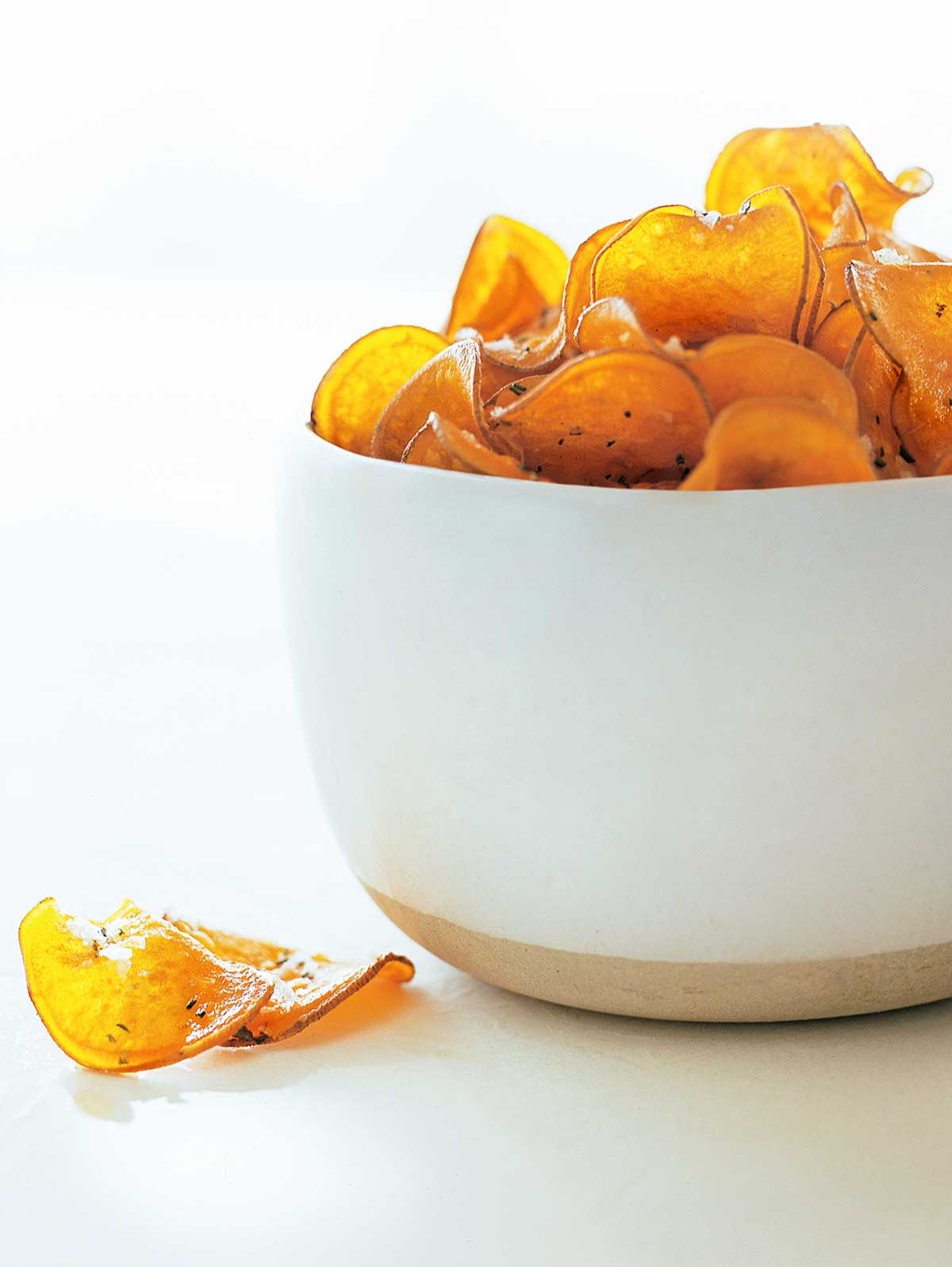 Sea salt-rosemary sweet potato chips piled in a white bowl, with a few laying beside it.