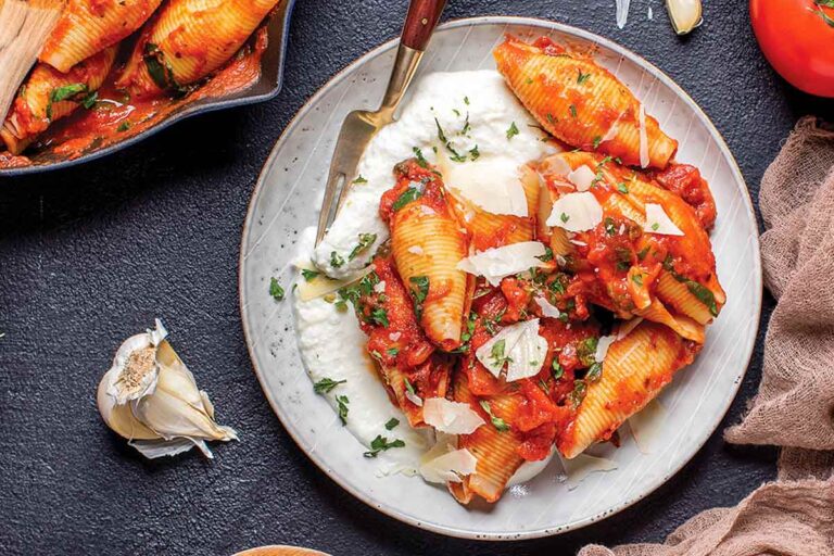 Shells with ricotta and fire-roasted tomato sauce on a white plate with a fork, beside a pan of shells, a clove of garlic and a tomato.