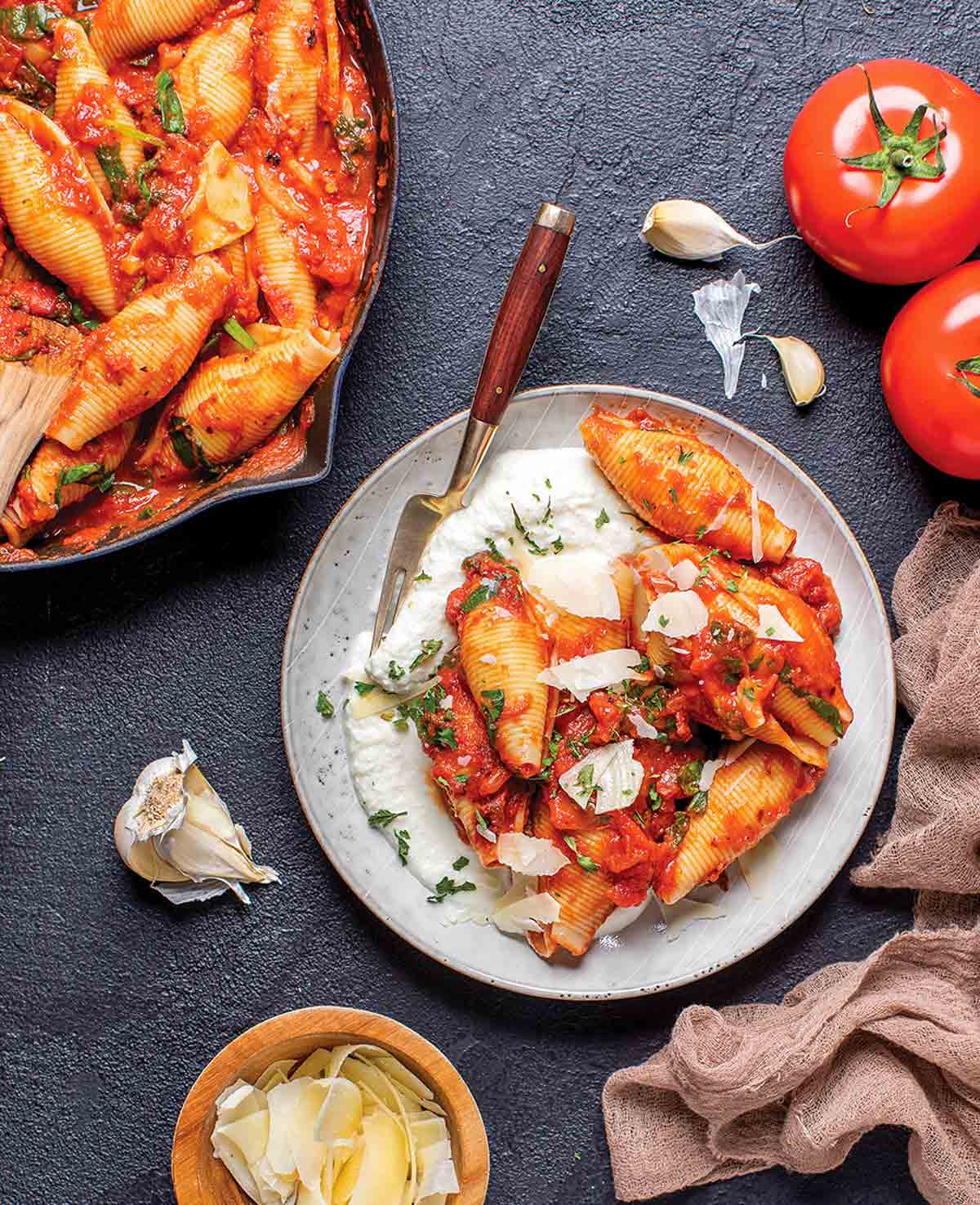 Skal med ricotta och eldrostad tomatsås på en vit tallrik med en gaffel, bredvid en panna med skal, en vitlöksklyfta, parmesan och en tomat.