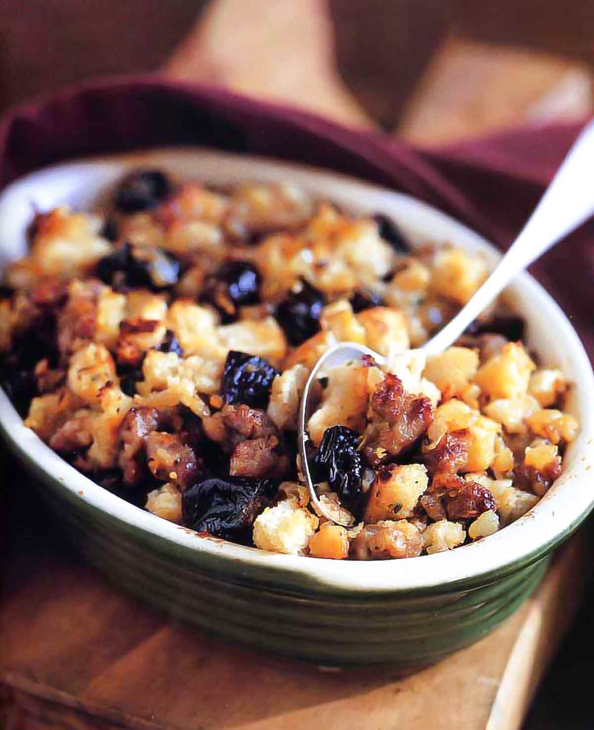 Sourdough dressing with sausage and prunes in a green ceramic dish with a large serving spoon.