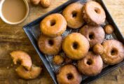 Spiced cider doughnuts and doughnut holes on a square metal tray with one in pieces beside a cup of coffee.