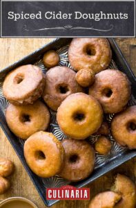 Spiced cider doughnuts and doughnut holes on a square metal tray with one in pieces beside a cup of coffee.