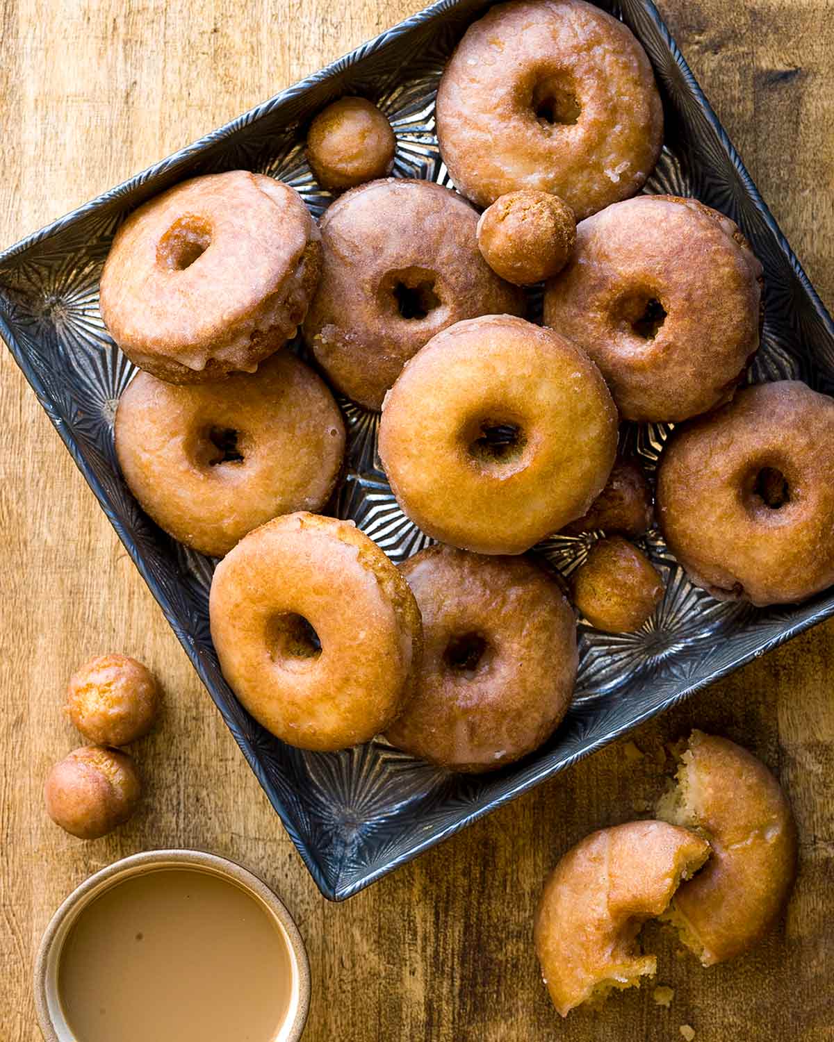 Spiced cider doughnuts and doughnut holes on a square metal tray with one in pieces beside a cup of coffee.