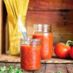 Spicy marinara sauce in a small mason jar with a spoon, with a larger mason jar, a jar of dry spaghetti, two tomatoes on the vine, and parsley in the background.