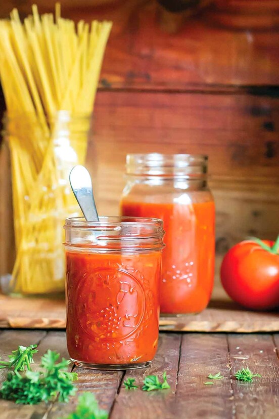 Spicy marinara sauce in a small mason jar with a spoon, with a larger mason jar, a jar of dry spaghetti, two tomatoes on the vine, and parsley in the background.