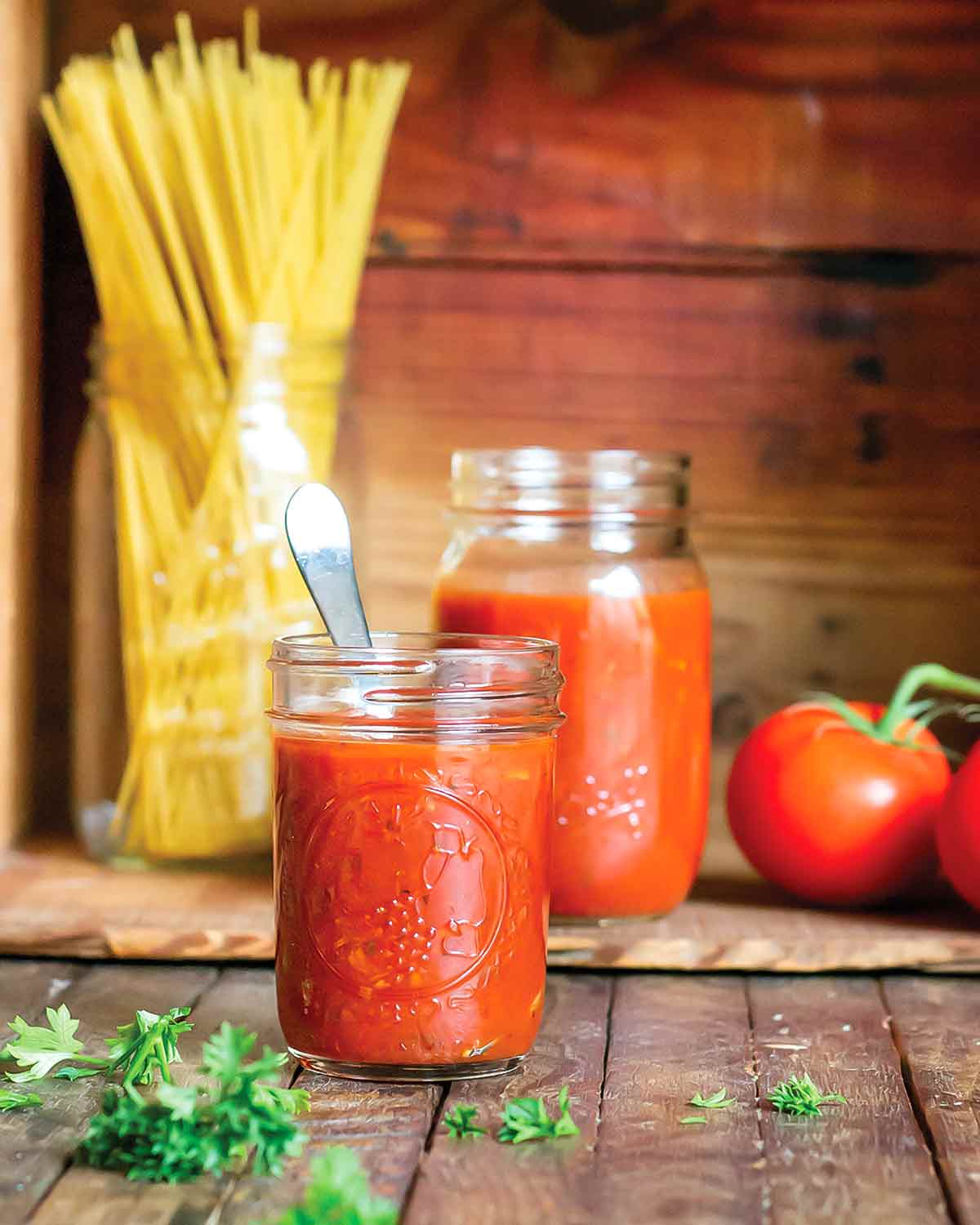 Spicy marinara sauce in a small mason jar with a spoon, with a larger mason jar, a jar of dry spaghetti, two tomatoes on the vine, and parsley in the background.