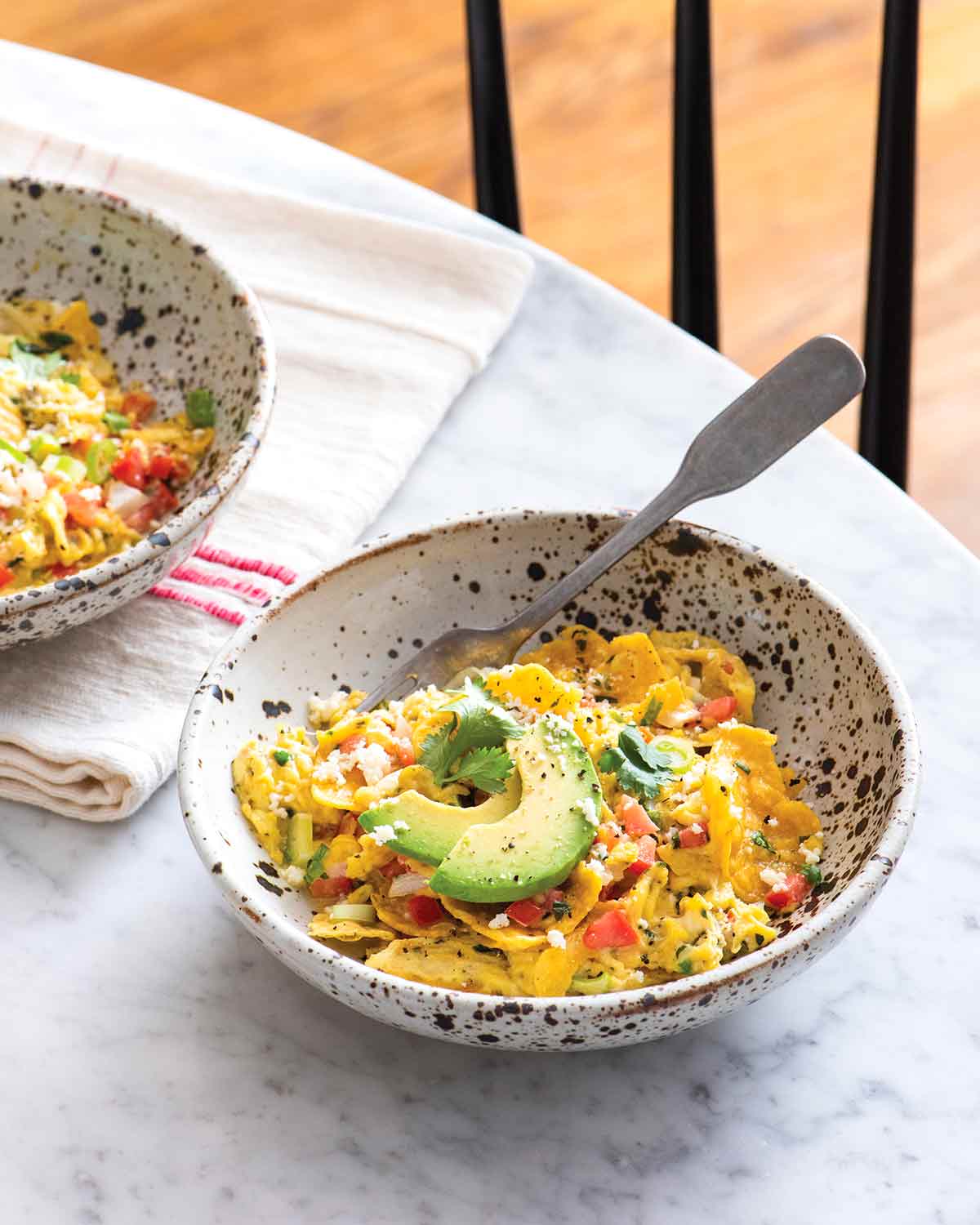 Tex-Mex migas topped with avocado in a white and blue bowl with a fork, beside a second bowl sitting on a tea towel.