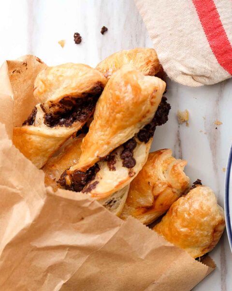 Torsades au chocolat in a brown paper bag on a marble counter, beside a white plate with pastry and a linen tea towel.
