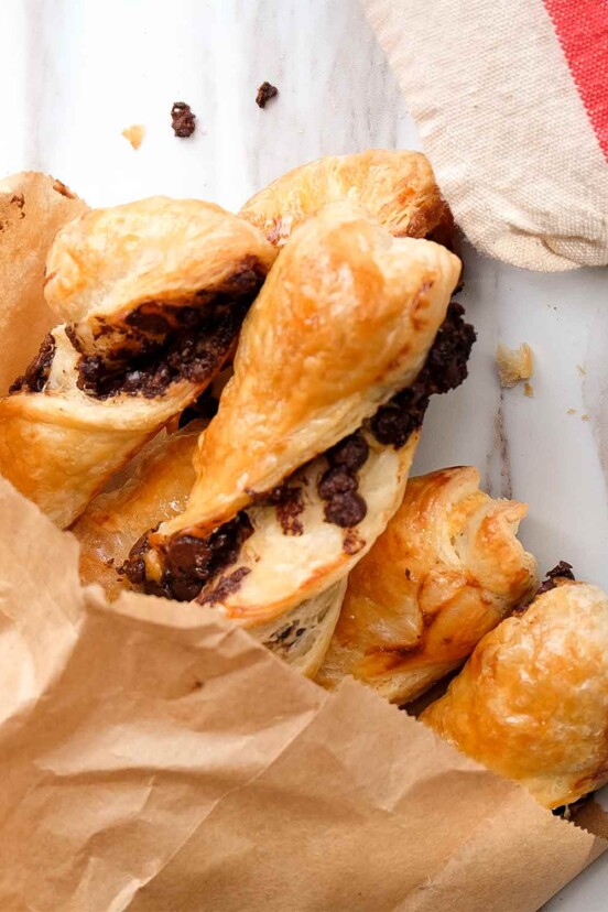 Torsades au chocolat in a brown paper bag on a marble counter, beside a white plate with pastry and a linen tea towel.