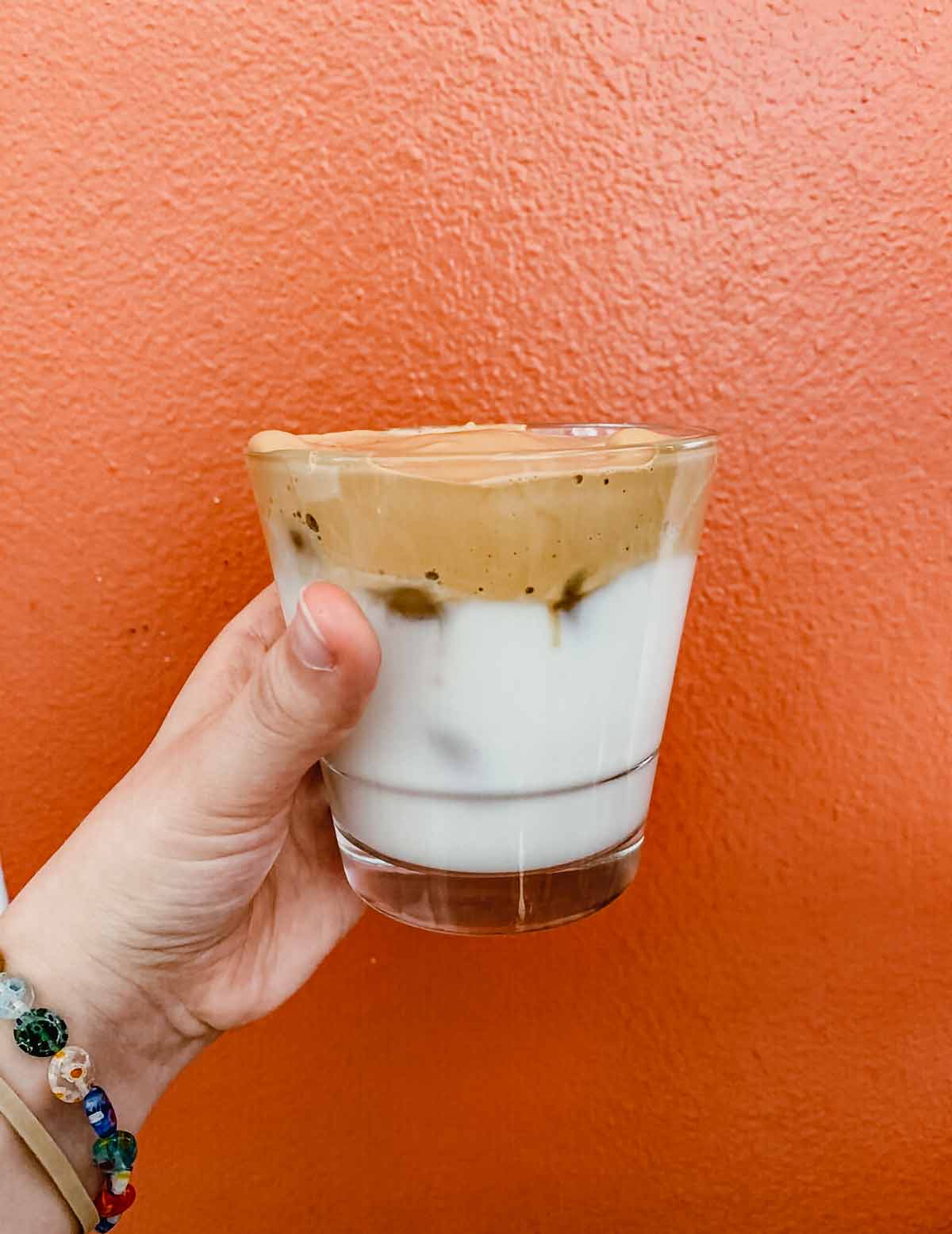 Whipped coffee in a clear glass, being held in front of an orange wall.