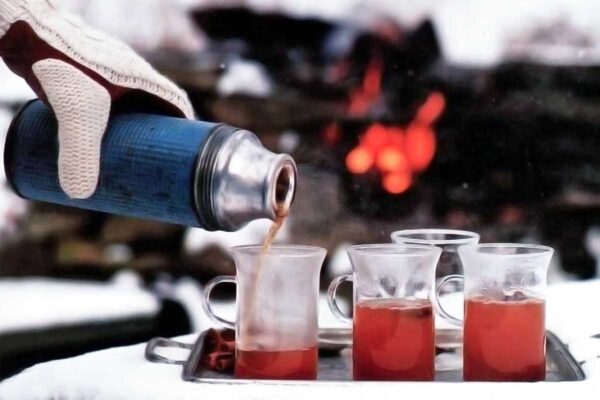 Apple spiced bourbon toddies on a tray in front of an outdoor fire, one being poured from a thermos in a gloved hand.