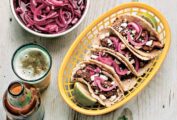 Four beef chili tacos and two lime halves in a basket, with bowls of pickled onions, slaw, and a beer on the side.