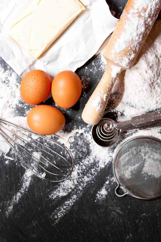 A stick of butter, three eggs, a whisk, a rolling pin, measuring spoons, a sieve, and flour on a black surface.