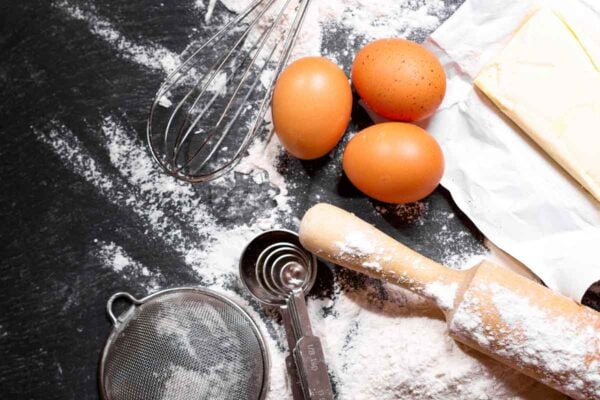 A stick of butter, three eggs, a whisk, a rolling pin, measuring spoons, a sieve, and flour on a black surface.