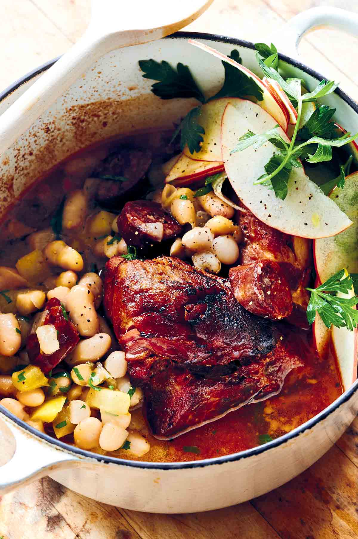 Braised pork shoulder with chorizo and white beans in a white enamel Dutch oven, garnished with apple salad.