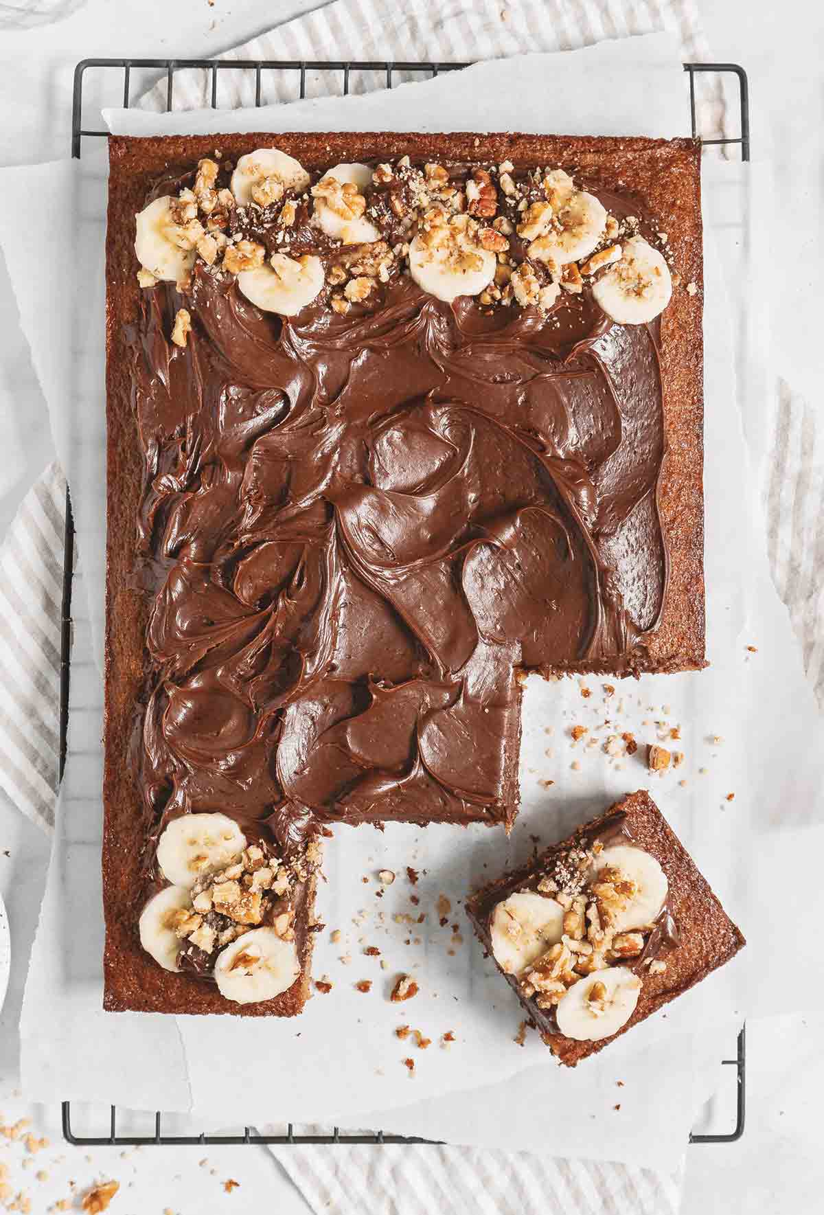 Brown butter banana bread sheet cake on a wire rack, with a few pieces cut out and garnished with bananas and chopped nuts.