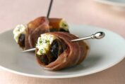 Two cheese-stuffed dates with Proscuitto on a white plate with metal pins securing them.