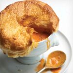 Clove-infused tomato soup in a white bowl, topped with a flaky pastry lid, on a plate with a spoon.