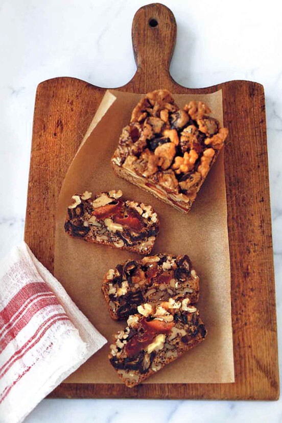 Dried fruit and nut cake in slices on a wooden cutting board with parchment paper, beside a red and white dish towel.