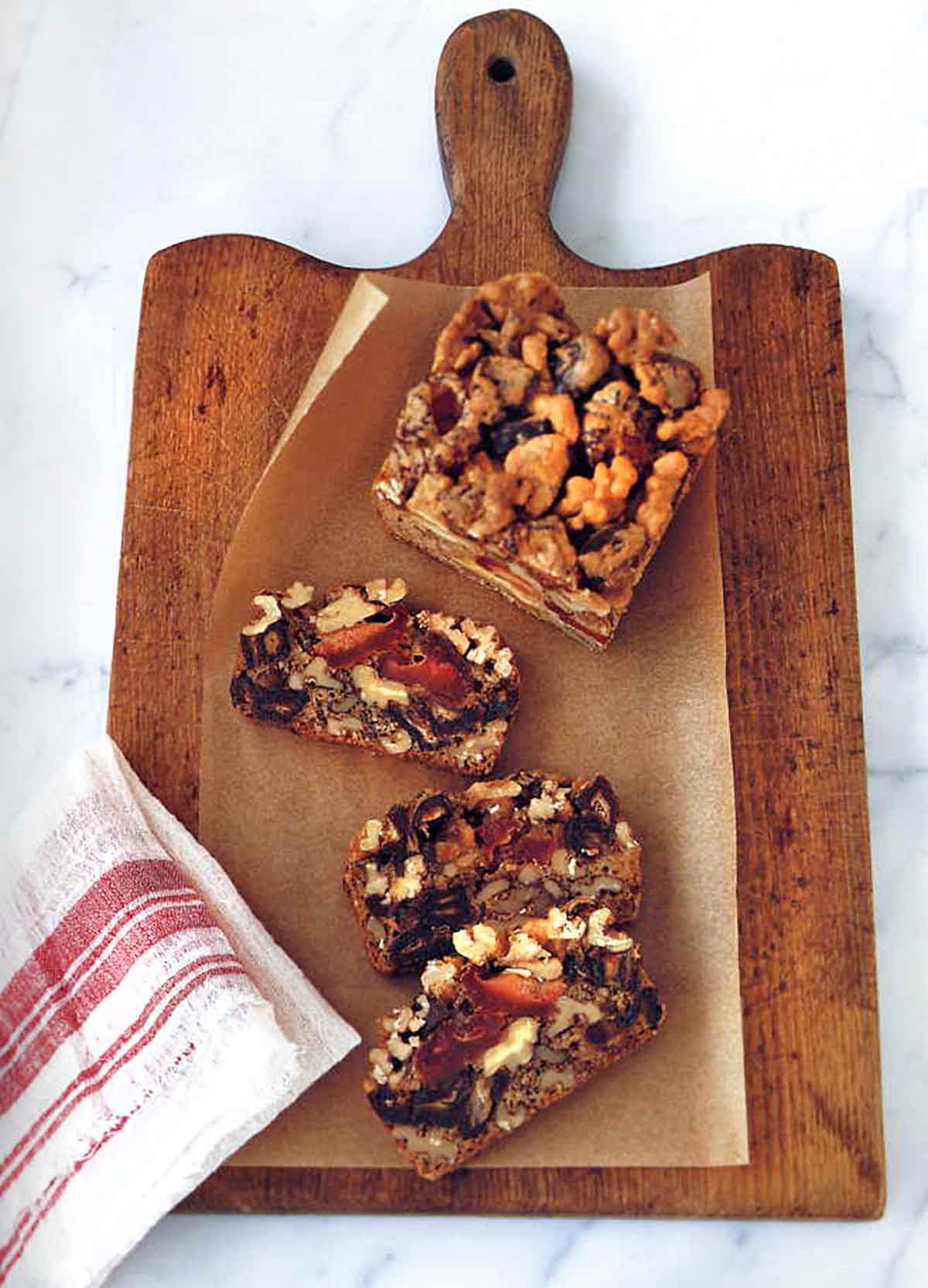 Dried fruit and nut cake in slices on a wooden cutting board with parchment paper, beside a red and white dish towel.