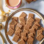 Gingerbread biscuits on a gold-rimmed plate, flanked by two cups of tea and gold Christmas ornaments.