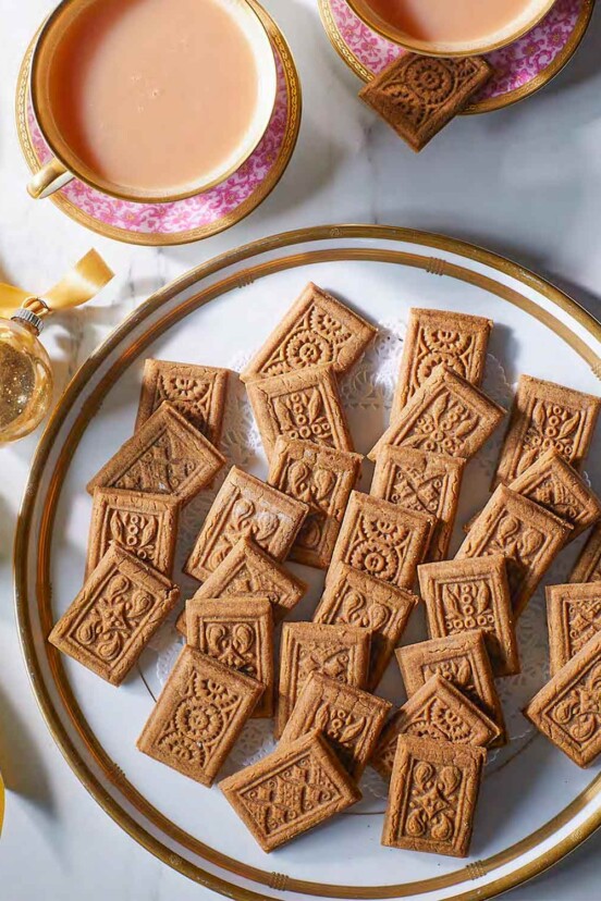 Gingerbread biscuits on a gold-rimmed plate, flanked by two cups of tea and gold Christmas ornaments.