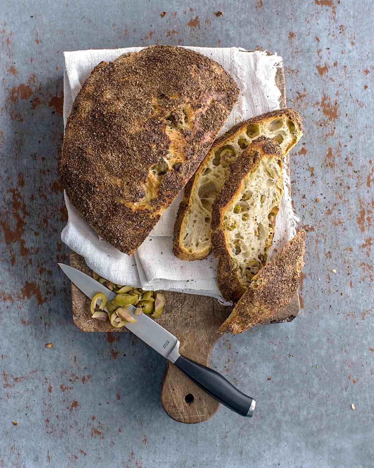 Jim Lahey no-knead olive bread with three slices cut, sitting on a wooden cutting board with a napkin, a bread knife, and a pile of sliced green olives.