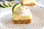 Key lime pie bars, cut into squares and garnished with whipped cream, lime zest, slices of lime, and chopped almonds.