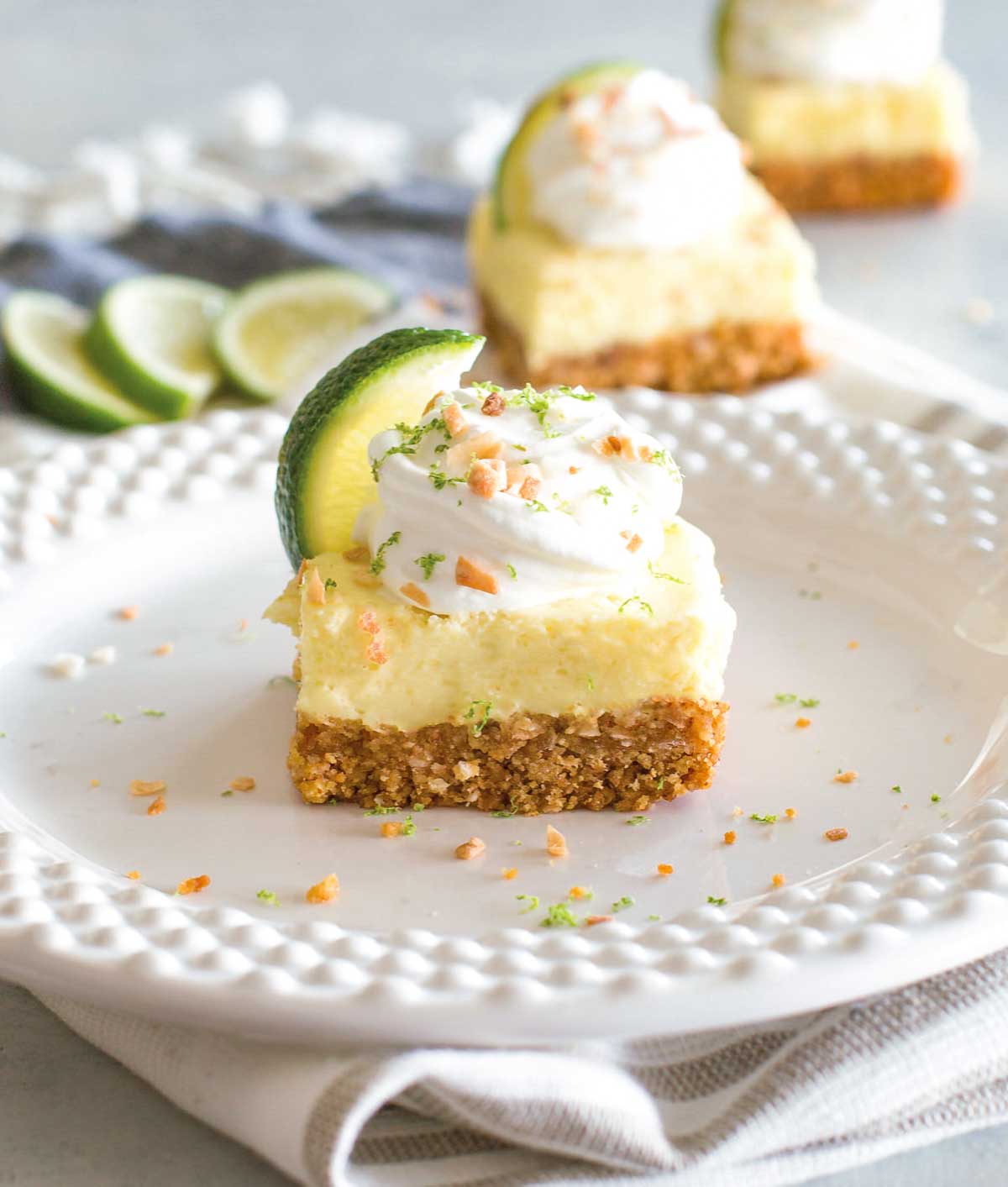 Key lime pie bars, cut into squares and garnished with whipped cream, lime zest, slices of lime, and chopped almonds.