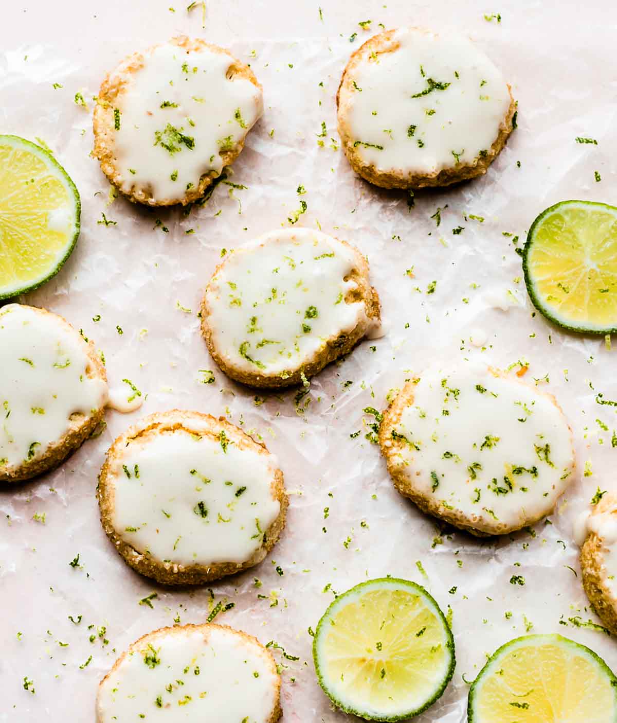 Key lime slice and bake cookies garnished with lime zest beside slices of lime, all on a sheet of parchment paper.