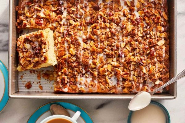 Make-ahead cinnamon coffee cake in a large rectangular pan, with two squares cut out, beside two cups of coffee and a bowl of drizzle with a spoon.