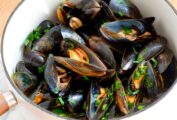 Mussels marinara garnished with parsley, in a white saucepan beside two wine glasses.