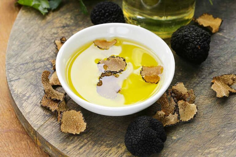 Truffle oil in a white bowl with dried truffle shavings floating in it, on a wooden cutting board with black truffles, truffle shavings, and a bottle of oil.