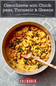 Orecchiette with chickpeas, turmeric, and greens in a large pot with a wooden spoon, on a countertop.