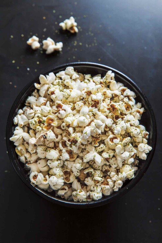 Popcorn with nori, citrus, and rosemary in a large black bowl, on a black background.